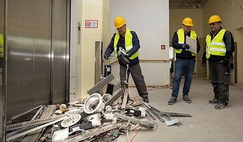 Ein Bauarbeiter des Kümmerei-Teams sortiert wieder verwendbare metallische Bauteile eines Abbruchgebäudes. Sein Teamleiter und ein Kollege stehen hinter ihm.