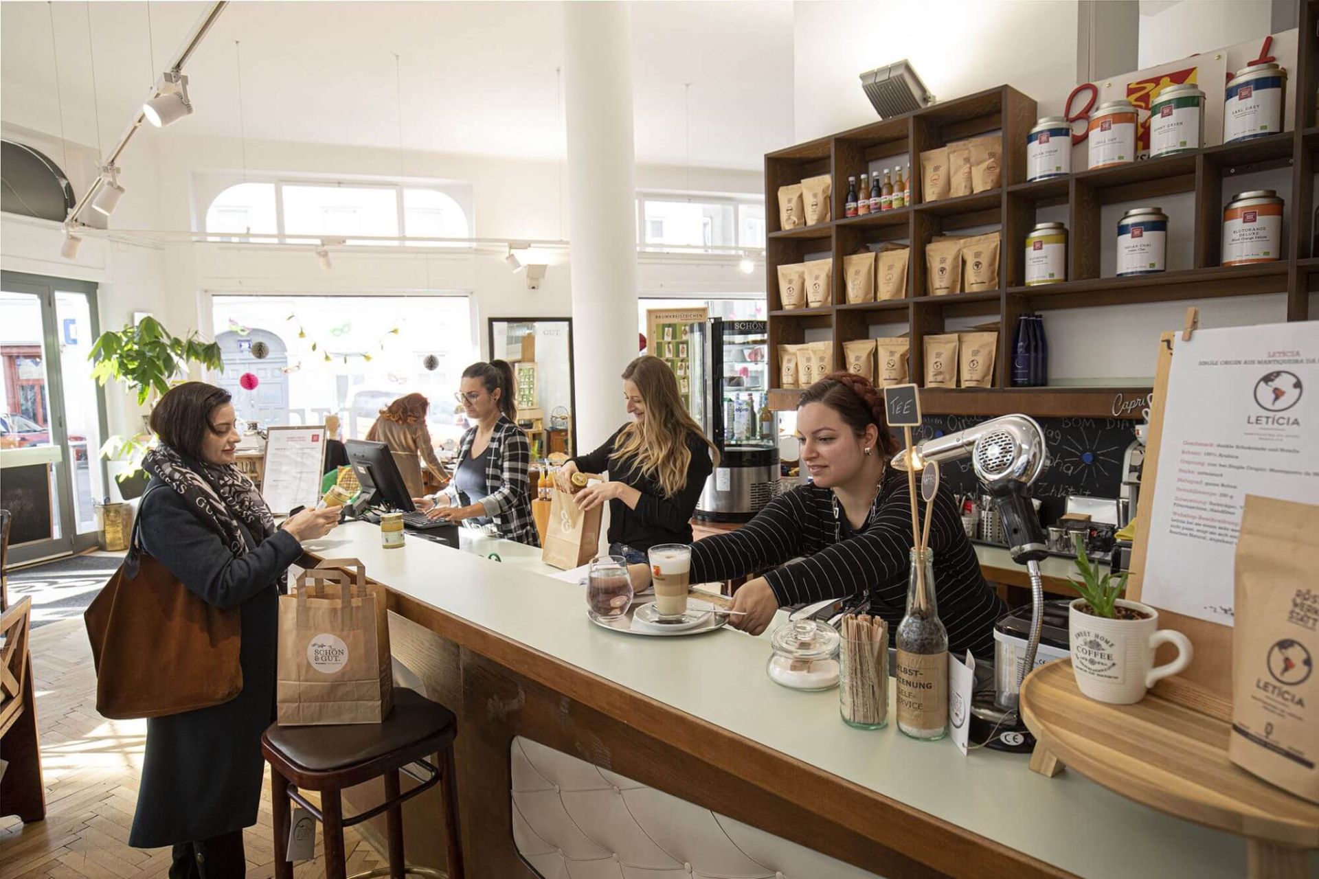 In unserem Social Concept Store Schön und Gut bereiten unsere Mitarbeiterinnen einen Kaffee und gekaufte Waren einer Kundin an der Theke vor.