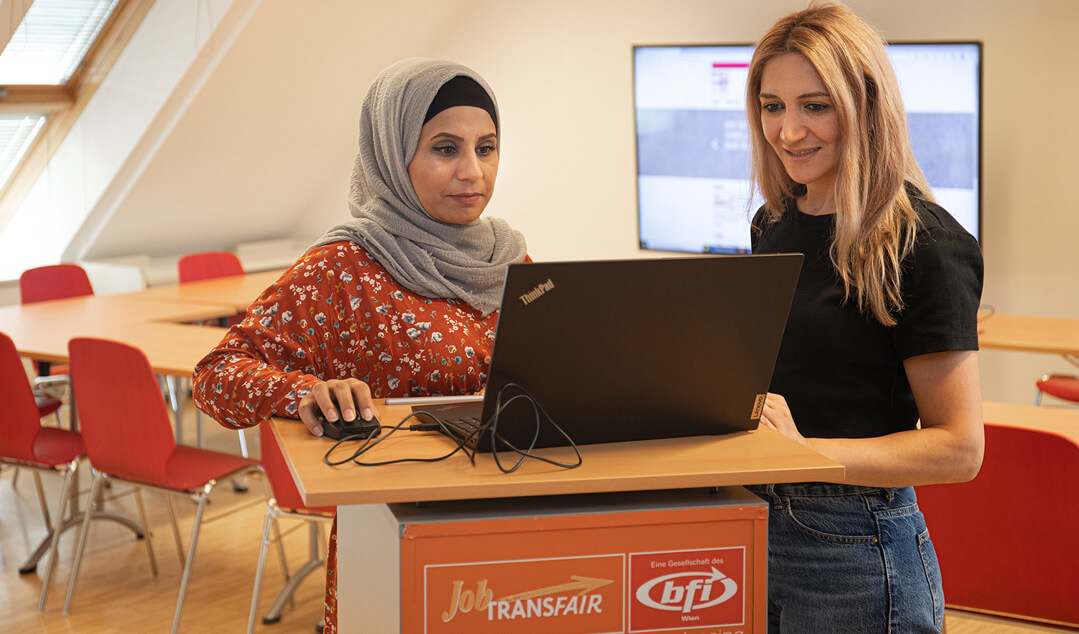 Zwei Damen stehen an einem orangen Job-TransFair-Pult, auf dem ein schwarzer Laptop steht. Beide schauen konzentriert auf den Bildschirm. Die Dame links bedient die Maus.