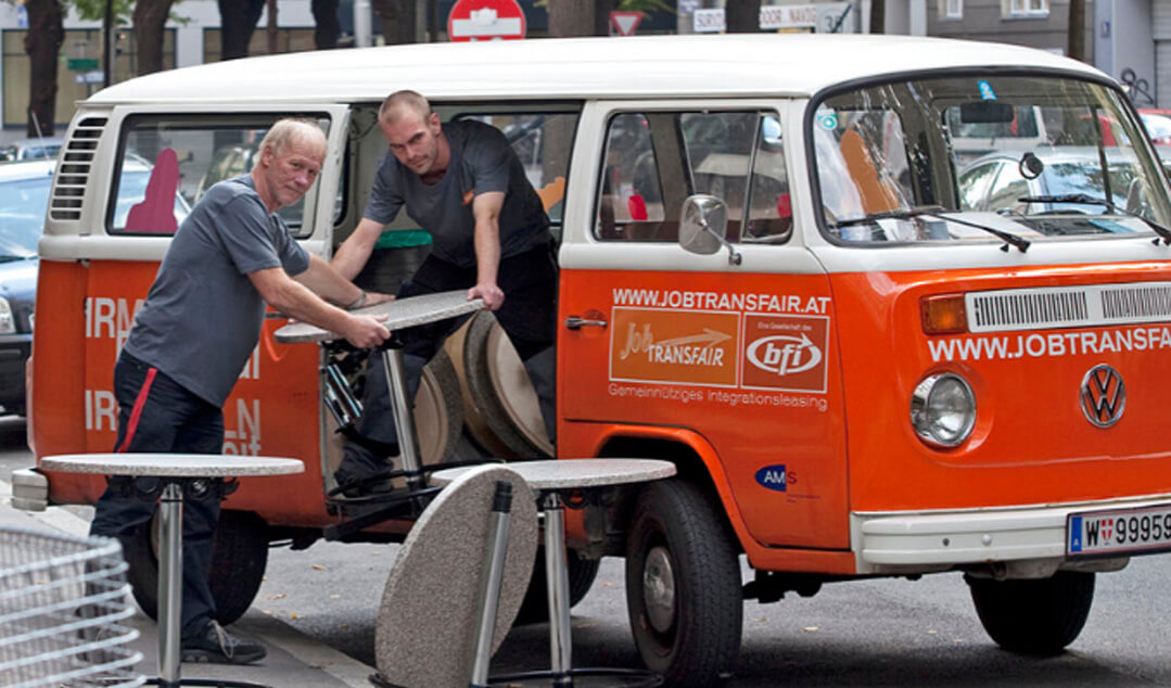Zwei Männer aus dem Kümmerei-Team entladen einen orangen Kleinbus mit Kaffeehaustischen. Sie halten inne und blicken direkt in die Kamera.