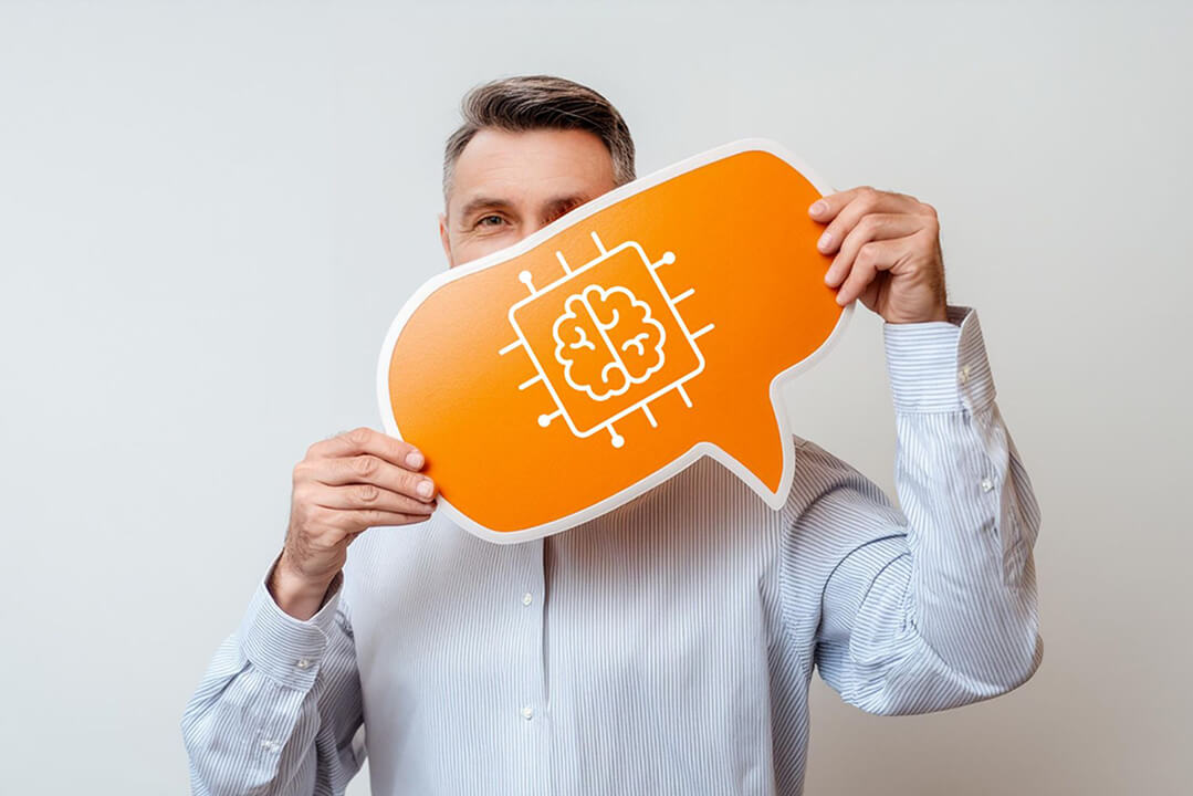 Ein Mann mittleren Alters mit einem blass gemusterten Hemd ist in halbnaher Perspektive zu sehen. Er hält ein oranges Schild, das die untere Hälfte seines Gesichts verdeckt. Das Schild zeigt das Piktogramm eines Gehirns in einem quadratischen Rahmen.