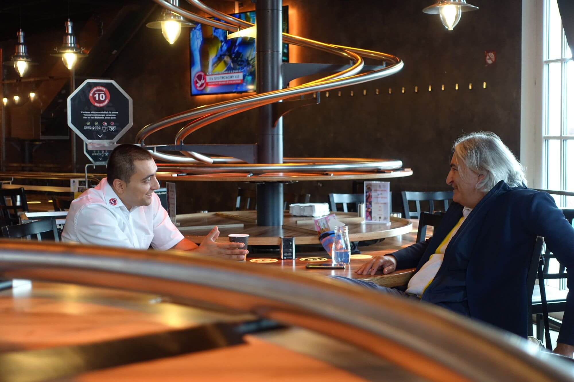 Martin Yakimov und Thomas Rihl an einem Tisch im Gespräch im Restaurant Rollercoaster. Im Vordergrund und Hintergrund die kleinen Achterbahnen.
